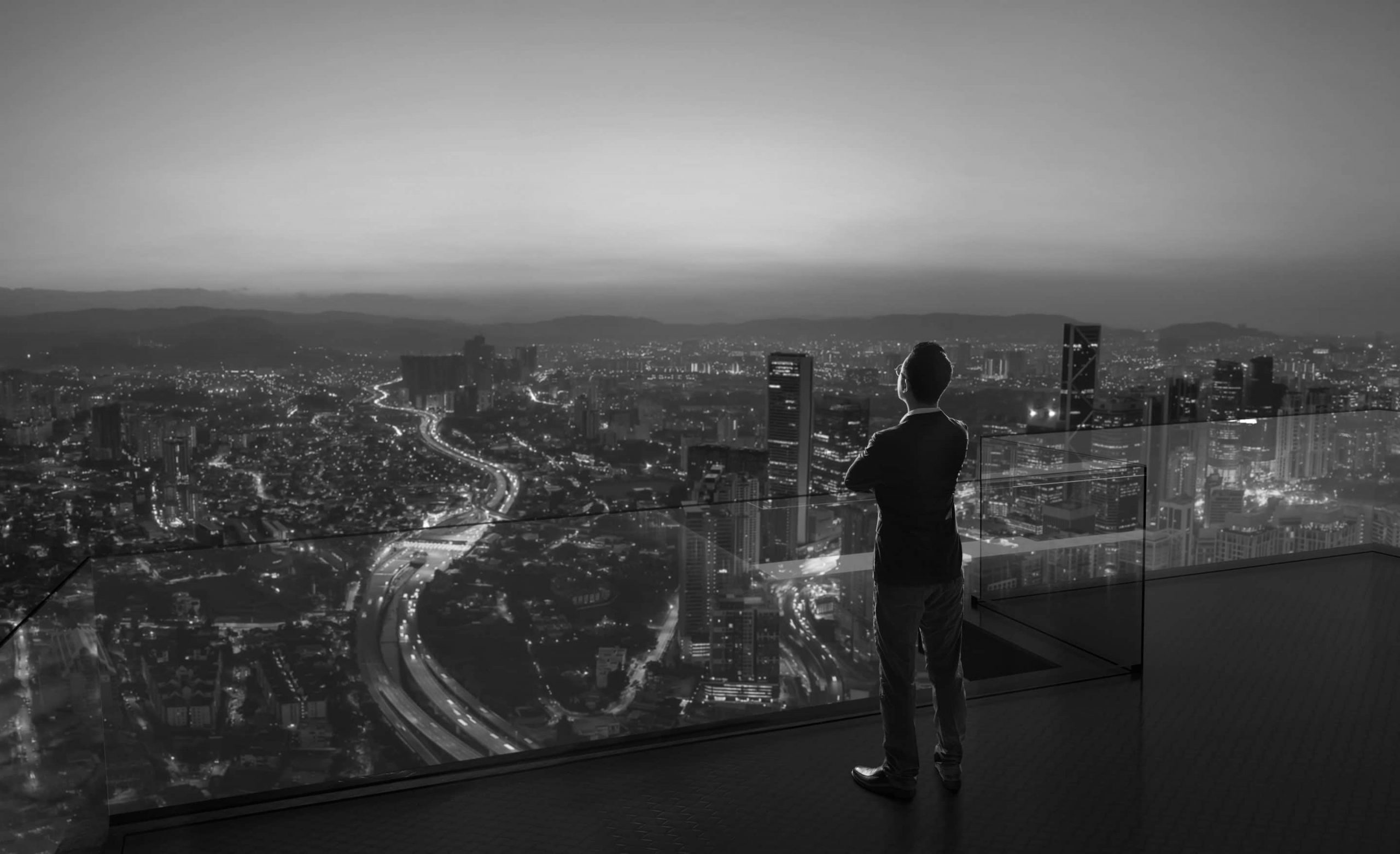 Businessman standing on open roof top balcony watching city night skyline