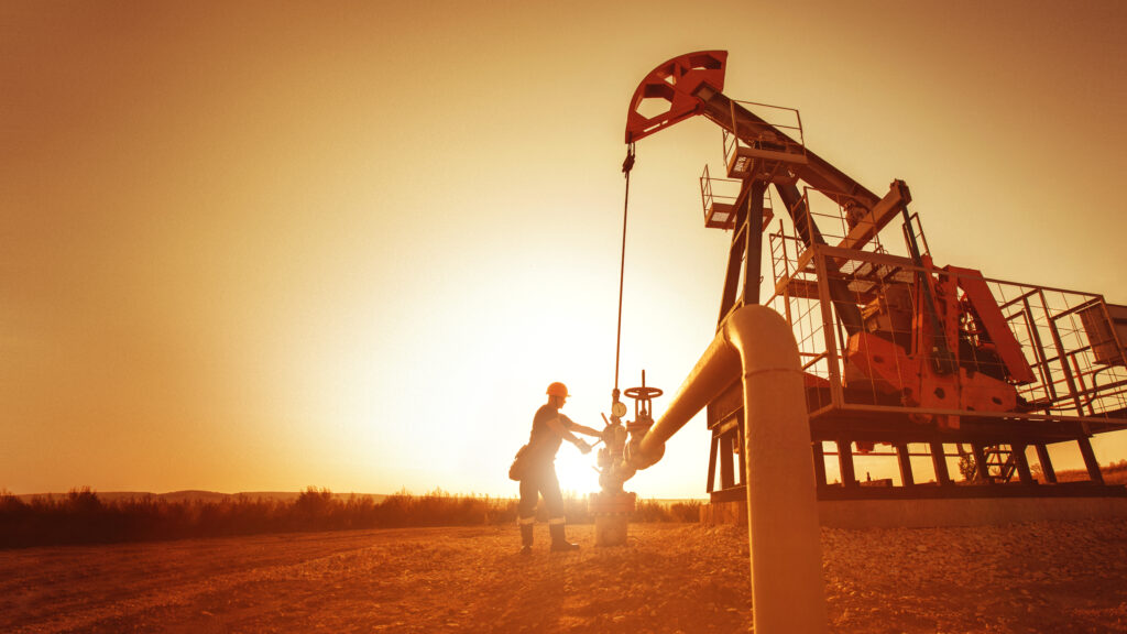 Oil field worker inspecting equipment beside a pumpjack at sunset.